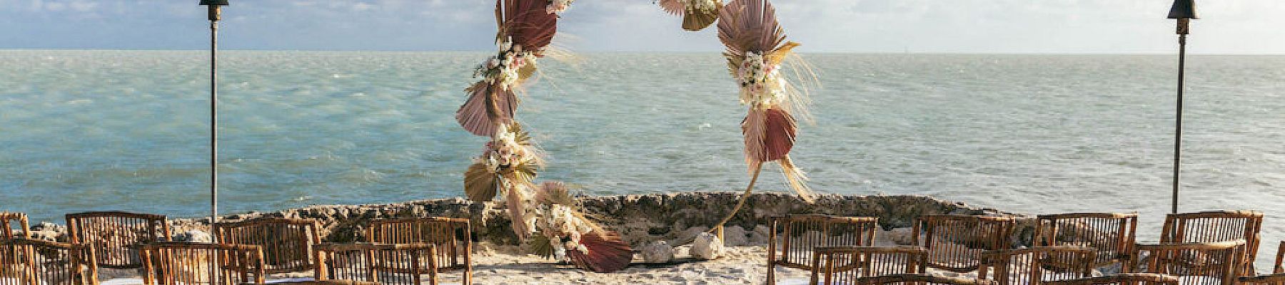 An outdoor wedding setup on a beach with rows of wooden chairs and a decorative arch facing the sea under a partly cloudy sky.