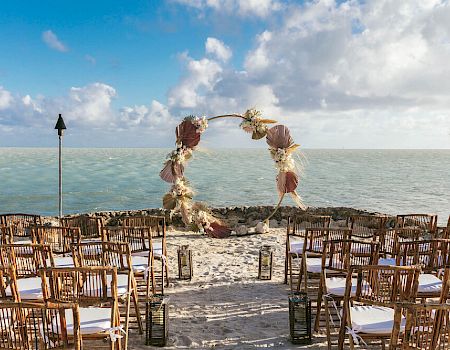 An outdoor wedding setup on a beach with rows of wooden chairs and a decorative arch facing the sea under a partly cloudy sky.