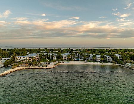 The image displays a coastal area with a small beachfront neighborhood, lush greenery, and calm waters under a partly cloudy sky.