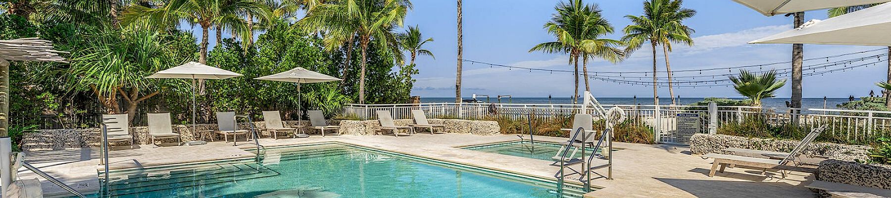 A serene tropical pool area with lounge chairs, umbrellas, and palm trees overlooking a water view under a clear blue sky.