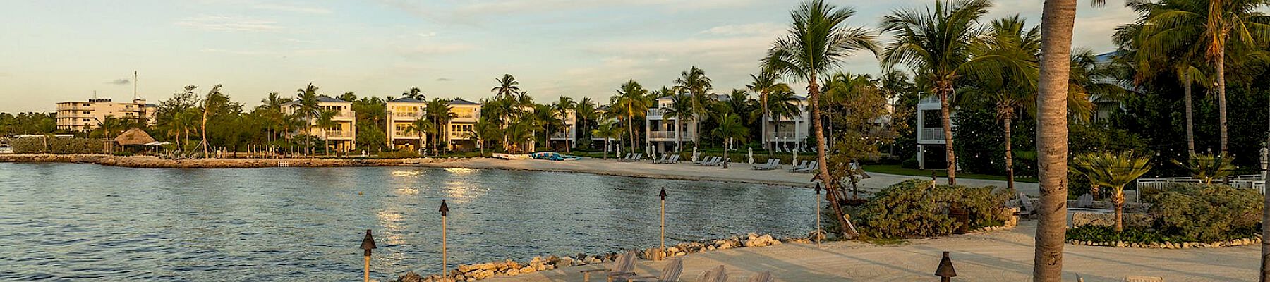 A tranquil beach scene with lounge chairs, palm trees, tiki torches, and a serene waterfront view in the background at sunset or dusk.