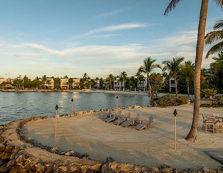 A tranquil beach scene with lounge chairs, palm trees, tiki torches, and a serene waterfront view in the background at sunset or dusk.