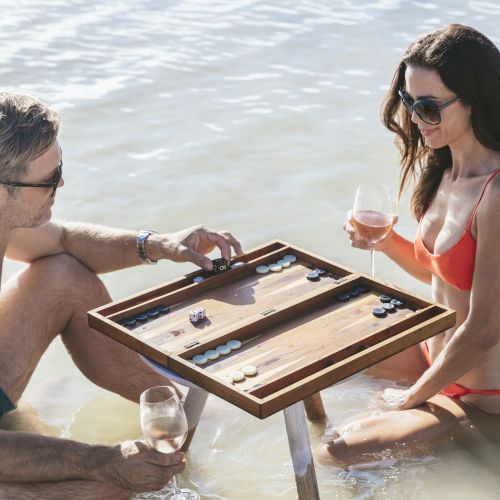 Two people are sitting in shallow water, playing a game of backgammon on a wooden board, while holding glasses of wine, both wearing swimsuits.