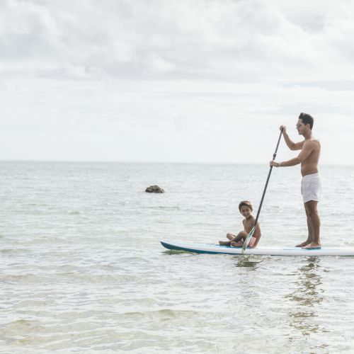 Two people are on a paddleboard in calm waters; the adult is standing while paddling, and the child is sitting relaxed, enjoying the ride.