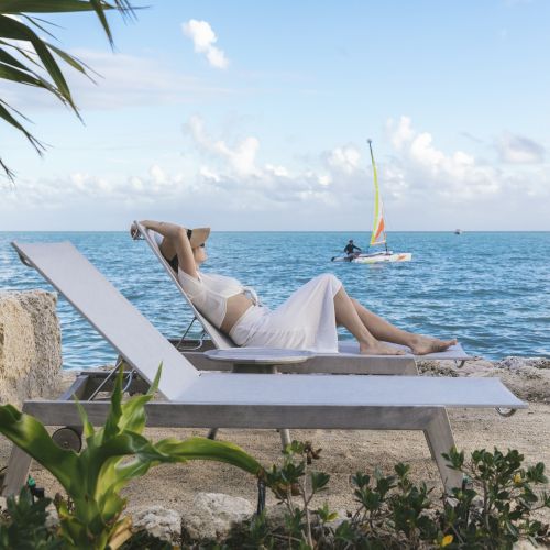 A person in a white outfit lounges on a chair by the sea, with a sailboat in the background and some greenery in the foreground.