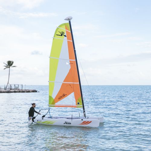 A person is sailing a small boat with colorful sails on a calm sea, with a few palm trees and a distant shore in the background.