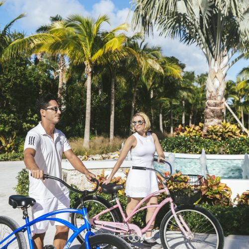 Two people in casual summer attire are with bicycles beside a tropical garden with palm trees and colorful plants, sky clear and the weather sunny.