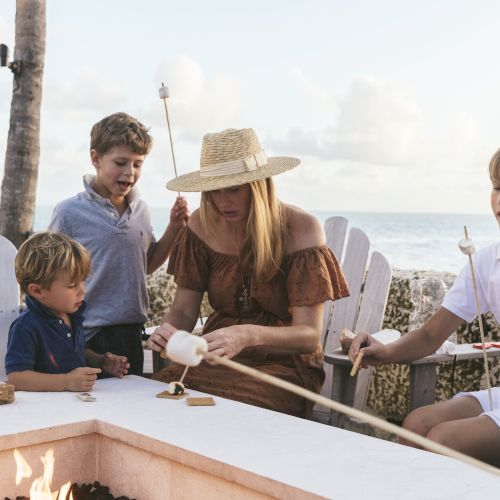 A family is toasting marshmallows around a fire pit. A woman and three children are seated outdoors by the beach, enjoying the activity.