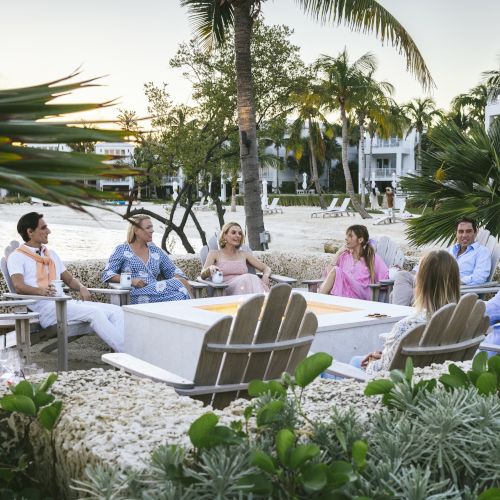 A group of people is sitting and conversing in a beachside setting, surrounded by lush greenery and palm trees.