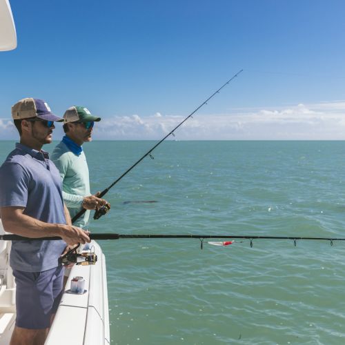 Two people fishing off the side of a boat in calm, blue waters under a clear sky.