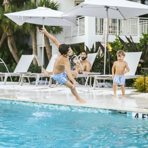 A boy jumps into a pool while a man and a younger boy, all in swimwear, look on; umbrellas and lounge chairs are in the background near greenery.