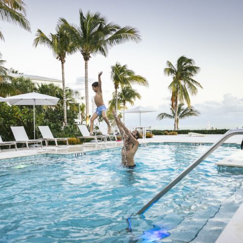 A man lifts a child in a pool against a backdrop of palm trees, lounge chairs, and umbrellas, creating a vibrant, tropical, and joyful atmosphere.