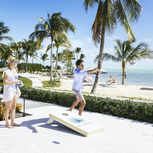 People playing cornhole on a seaside patio; palm trees, white sand, and blue ocean in the background.