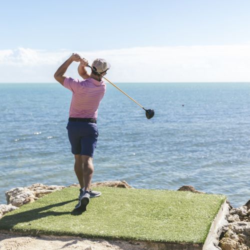 A golfer in a pink shirt and blue shorts swings a golf club on a small artificial green by the ocean, with clear blue skies in the background.