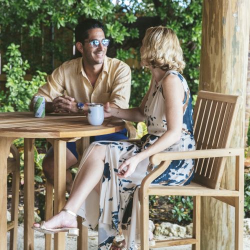 A man and a woman sitting at a wooden table under a thatched roof, surrounded by greenery, enjoying beverages.