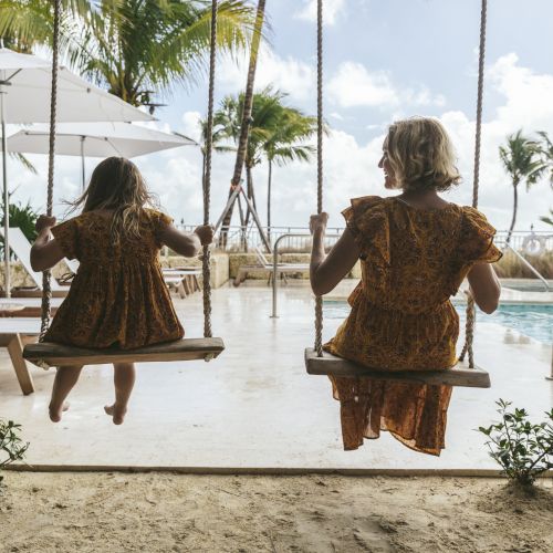 Two people are on swings by a pool, surrounded by palm trees and lounge chairs, under a sunny sky.