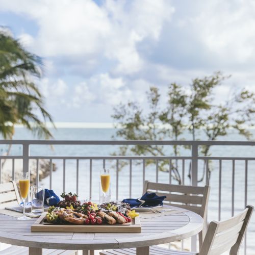 A balcony with a white table set for dining, featuring seafood, drinks, and a view of the ocean with palm trees and clouds.