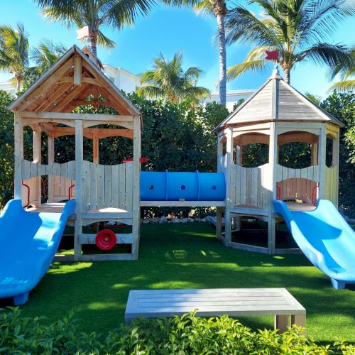 A playground with two wooden structures, blue slides, and a connecting tunnel is set in a sunny area with palm trees and green grass.