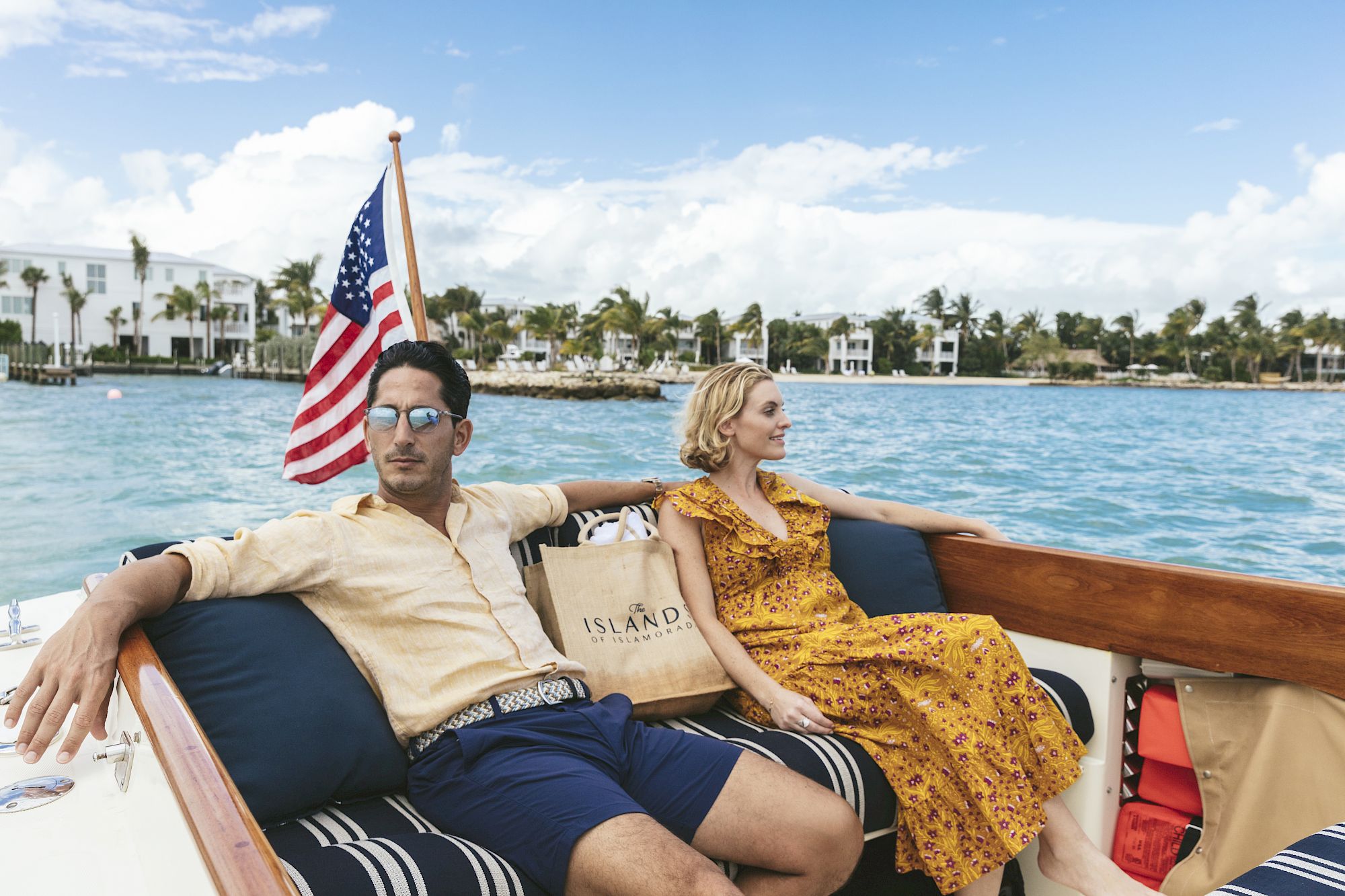 A man and woman are sitting on a boat, enjoying a sunny day on the water with an American flag in the background, and tropical scenery surrounds them.