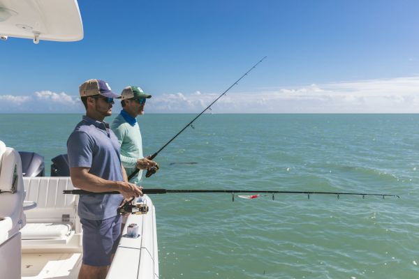 Two people are fishing off the side of a boat in clear, blue waters under a bright sky, with fishing rods in hand.