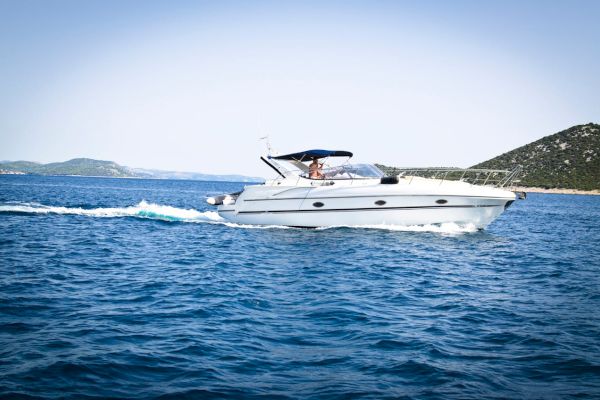 The image shows a white motorboat with a blue canopy cruising on a body of water, with distant islands and a clear sky in the background.