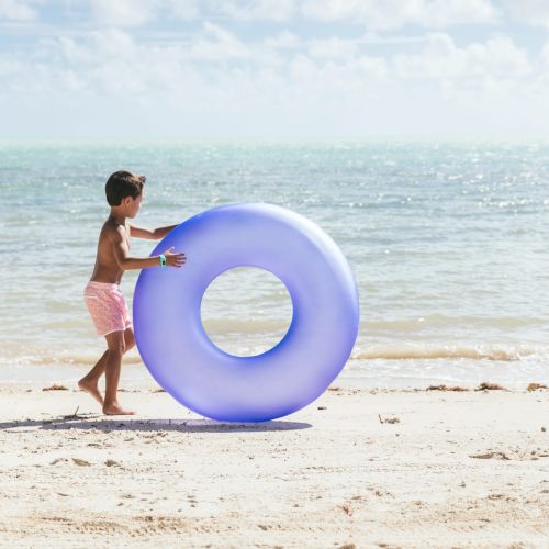 A child in pink shorts is pushing a large blue inflatable ring on a sandy beach with ocean waves in the background.