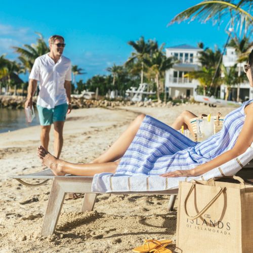 A person relaxes on a beach chair while another approaches, set against a backdrop of palm trees, blue skies, and beachfront houses.