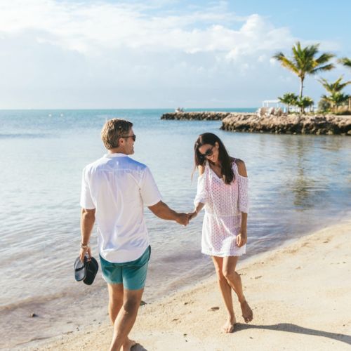 A couple walks hand in hand along a sandy beach by the ocean, with palm trees in the background on a bright, sunny day.