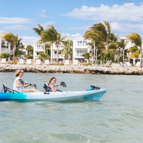 Two people are kayaking in the ocean with a coastal residential area featuring white buildings and palm trees in the background, ending the sentence.