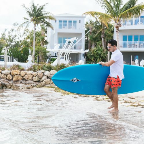 A man in red swim trunks is carrying a blue surfboard at the water's edge in front of beachfront houses and palm trees.