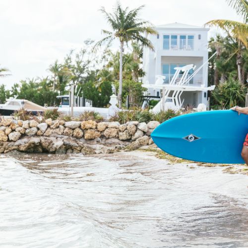 A person in red shorts is carrying a blue surfboard, walking towards the water on a beach with houses and palm trees in the background.