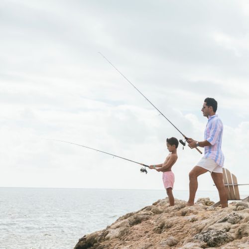 Two people are fishing off a rocky shore, standing close together, and casting their lines into the water.