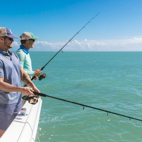 Two people are standing on a boat holding fishing rods and fishing in a calm, blue sea under a clear sky.