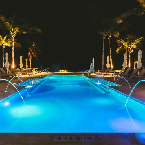 A well-lit swimming pool at night, surrounded by lounge chairs and palm trees, with fountains creating arcs of water over the pool.