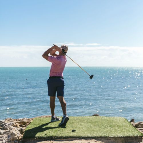 A person is swinging a golf club on a coastal golf course with the ocean in the background under a clear blue sky.