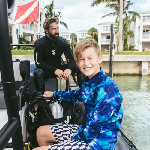 A smiling boy and a man in a wetsuit are on a boat near water with buildings and palm trees in the background, ready for a diving adventure.