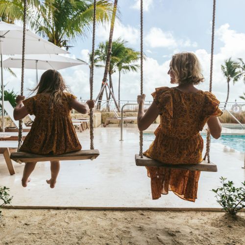 Two people wearing similar dresses are sitting on swings by a pool, surrounded by palm trees and lounge chairs, enjoying a sunny day.