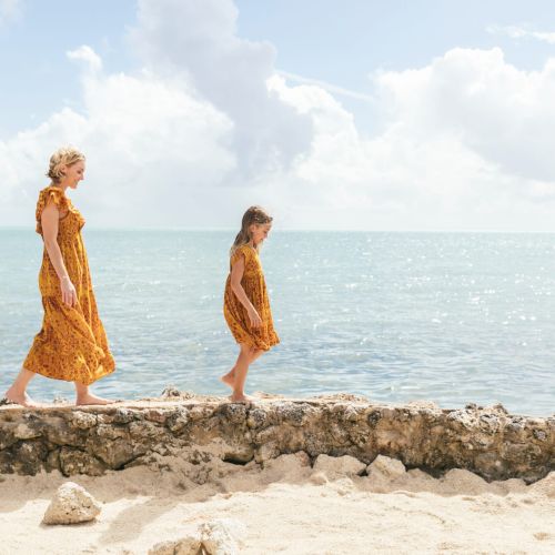 A woman and a girl in matching yellow dresses walk along a rocky shore by the ocean, under a partly cloudy sky.