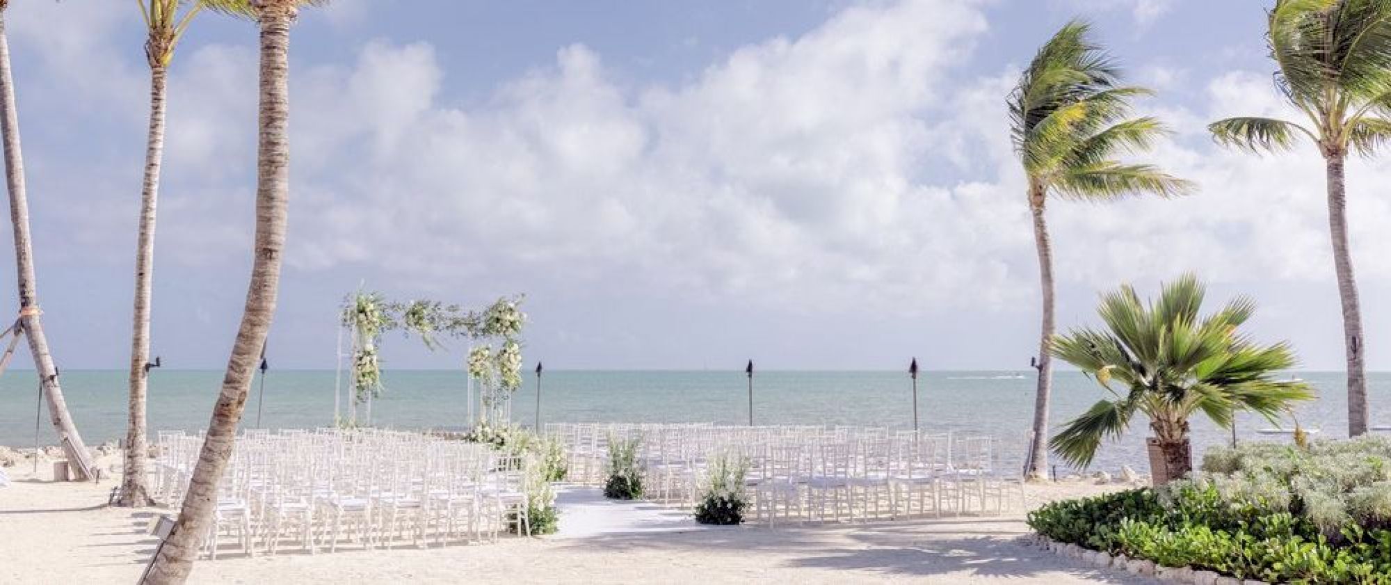 A beachside wedding setup with chairs arranged facing an altar on a sandy shore, surrounded by palm trees under a partly cloudy sky.