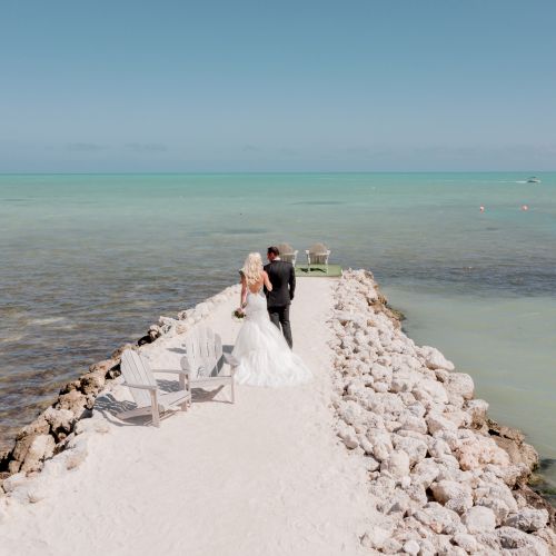 A couple stands on a rocky pier extending into a calm sea, with chairs on either side; the sky is clear and bright.