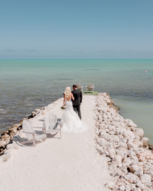 A couple stands on a rocky pier extending into a calm sea, with chairs on either side; the sky is clear and bright.