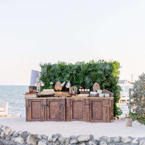 An outdoor setup with a wooden bar counter, lush greenery, and a view of the sea in the background.