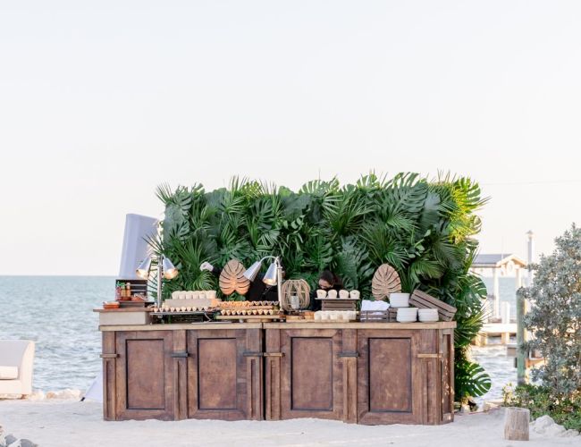 An outdoor setup with a wooden bar counter, lush greenery, and a view of the sea in the background.