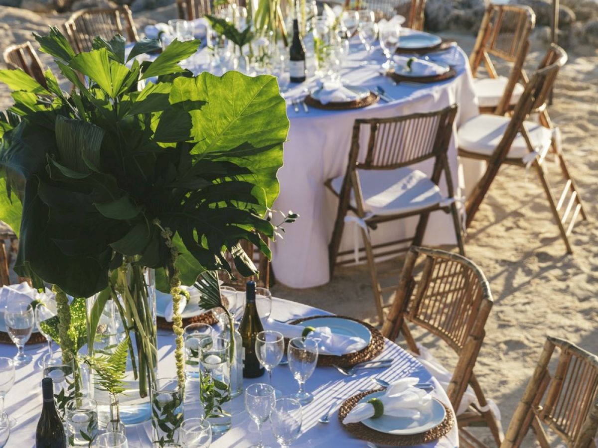 An elegant outdoor dining setup by the water with round tables adorned with large green centerpieces, white tablecloths, and wooden chairs.