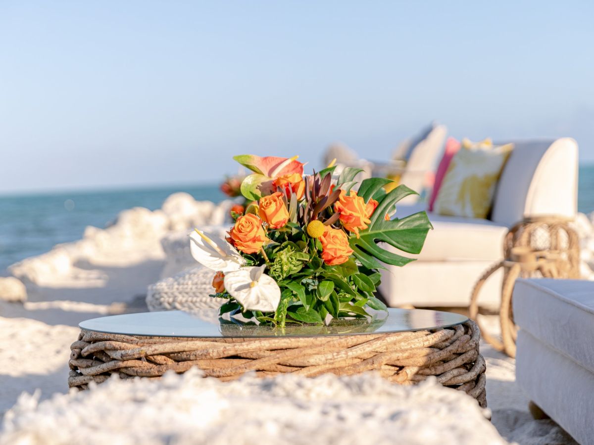 A bouquet of flowers sits on a wicker table on a sandy beach, with the ocean and white furniture in the background.