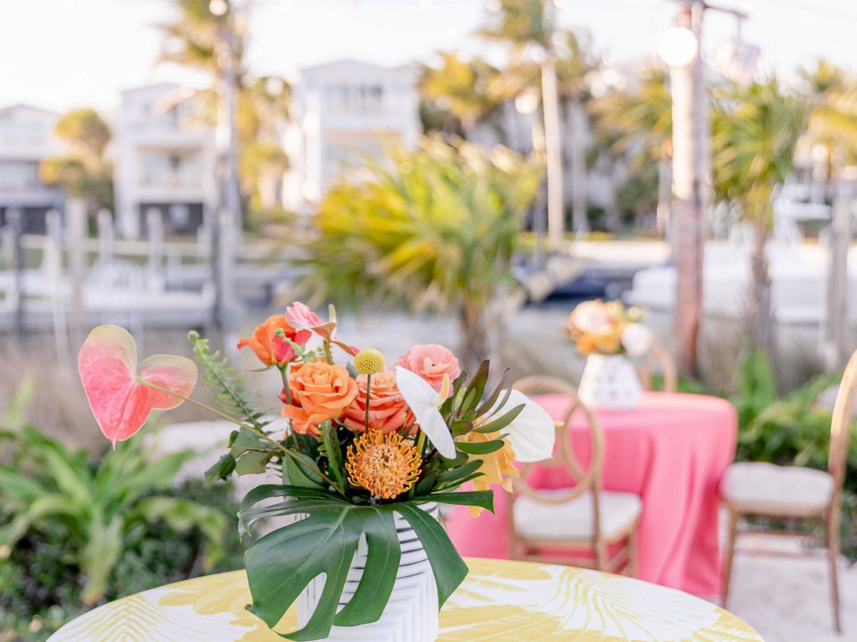 A tropical outdoor setting with a table covered in a yellow and white cloth, topped with a floral arrangement. Palm trees and buildings in the background.