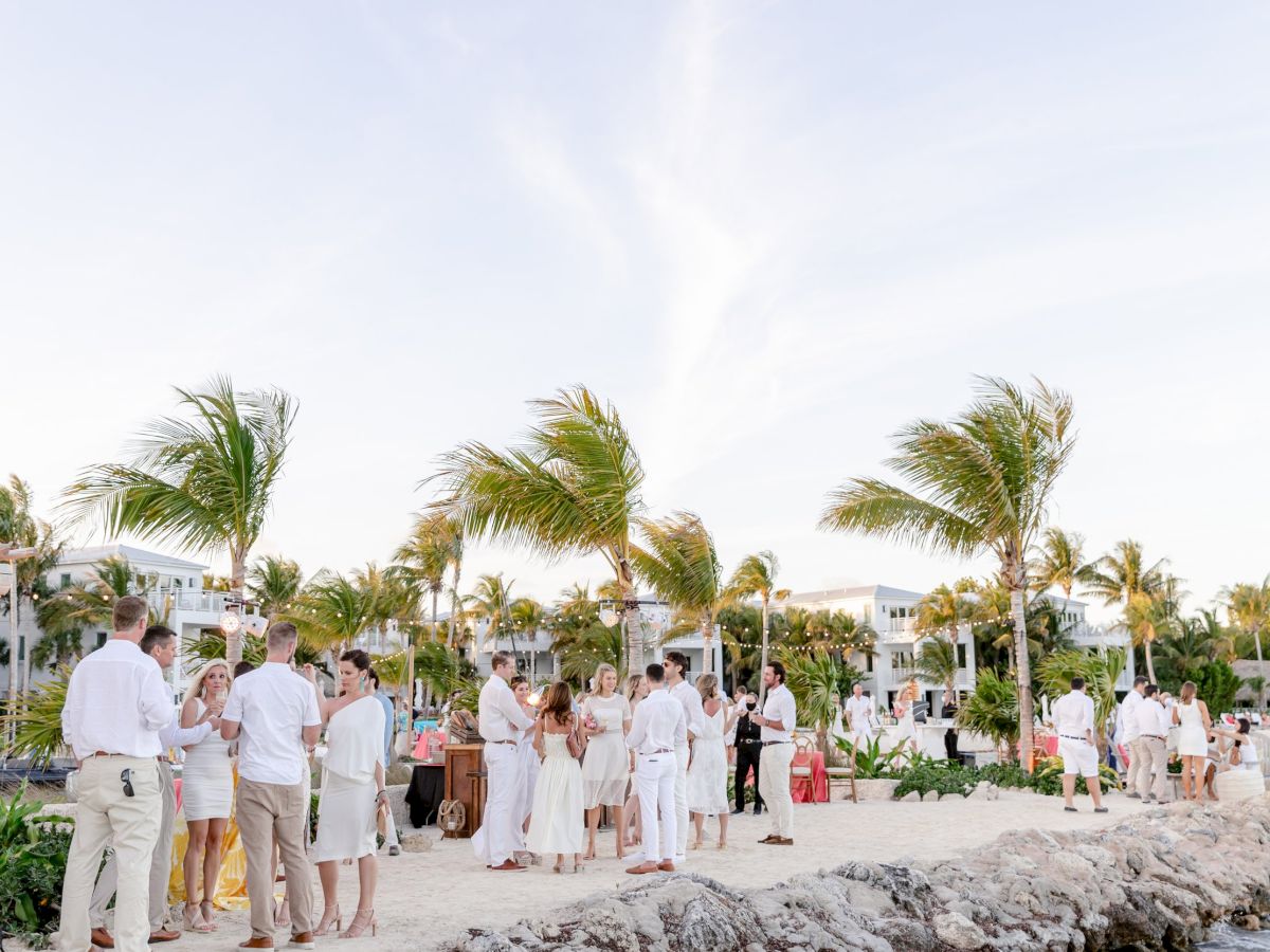 People dressed in white are gathered on a beach with palm trees and buildings in the background, enjoying a social event.