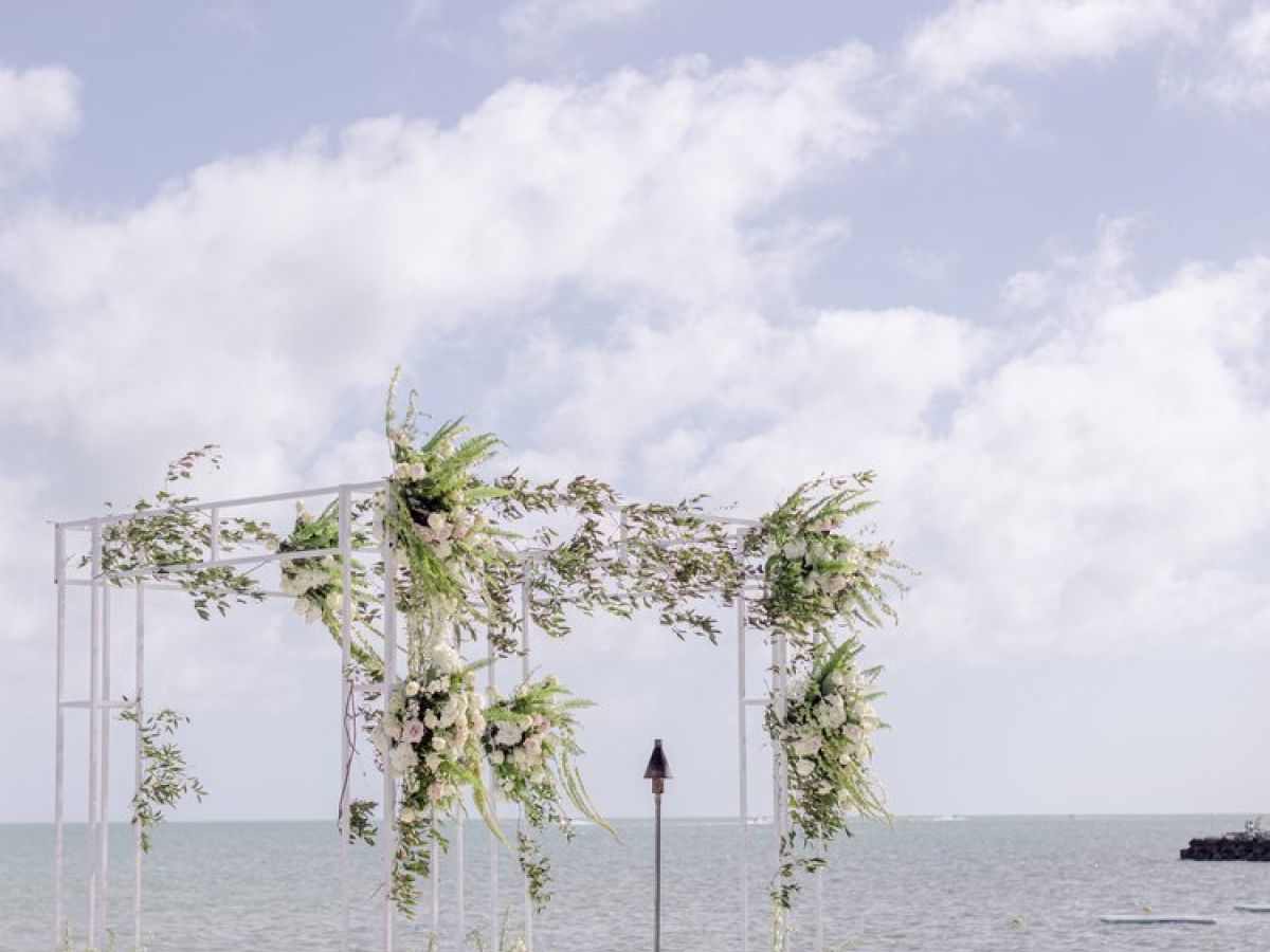 A seaside wedding setup with white chairs and a floral arch facing the ocean and puffy clouds in the sky.