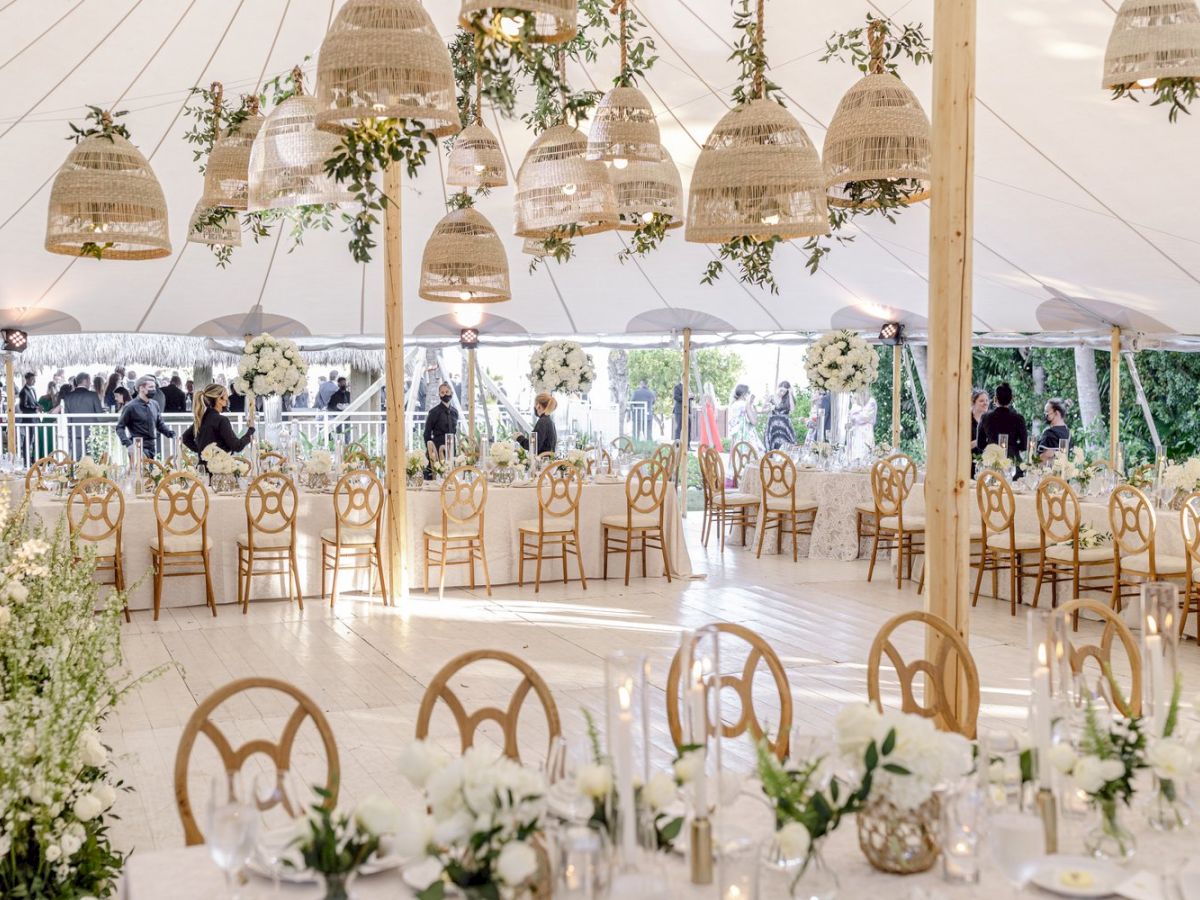 An elegant outdoor wedding reception setup under a tent, featuring wooden chairs, floral arrangements, and hanging lanterns.