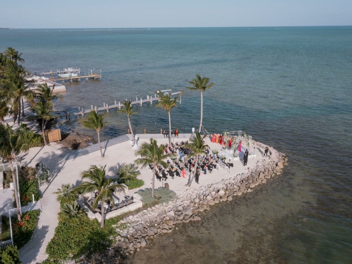 A beachfront with a rocky shoreline, palm trees, and people gathered on a small peninsula. The ocean stretches into the horizon.